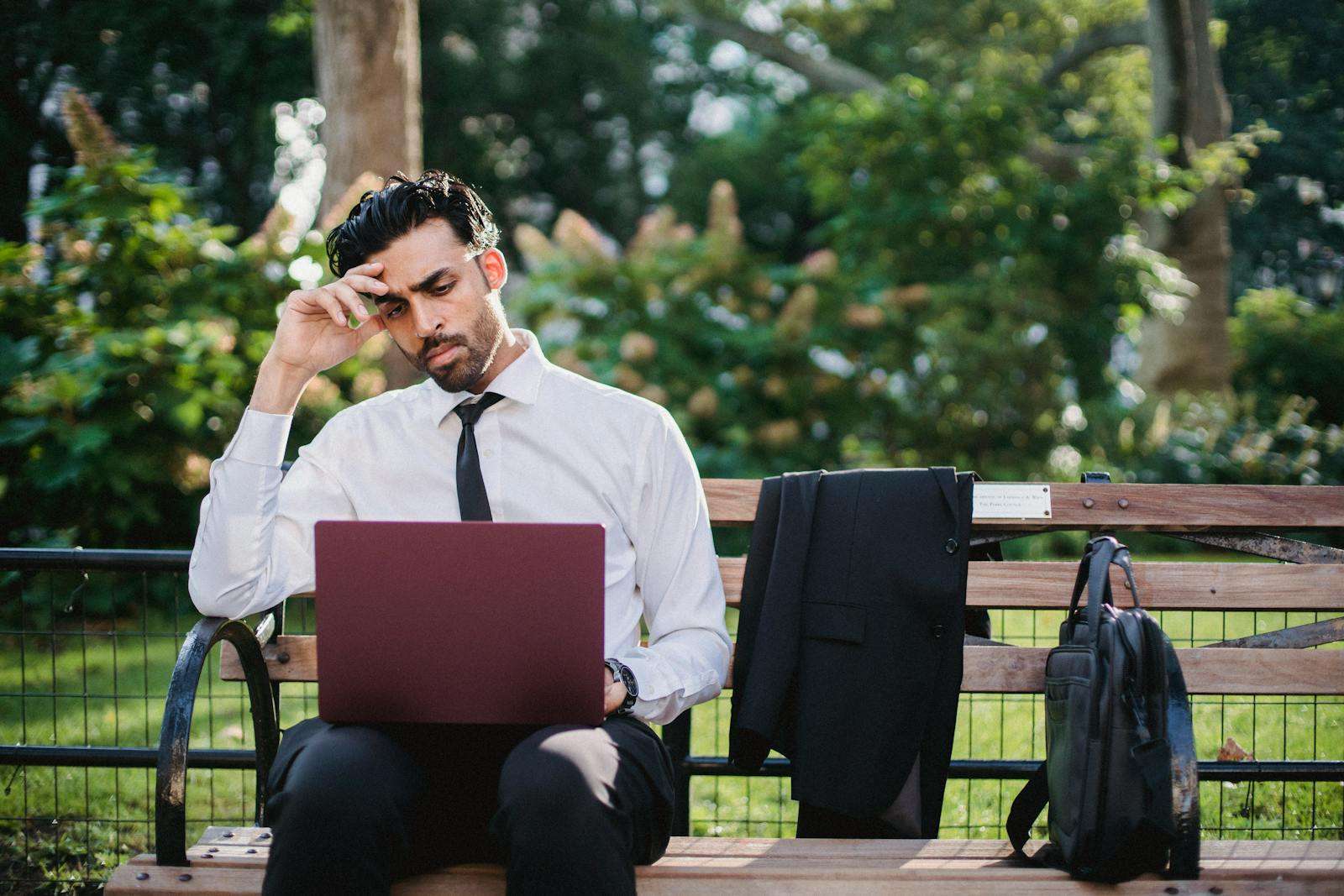 A thoughtful businessman works on his laptop while sitting on a park bench with his coat and bag.